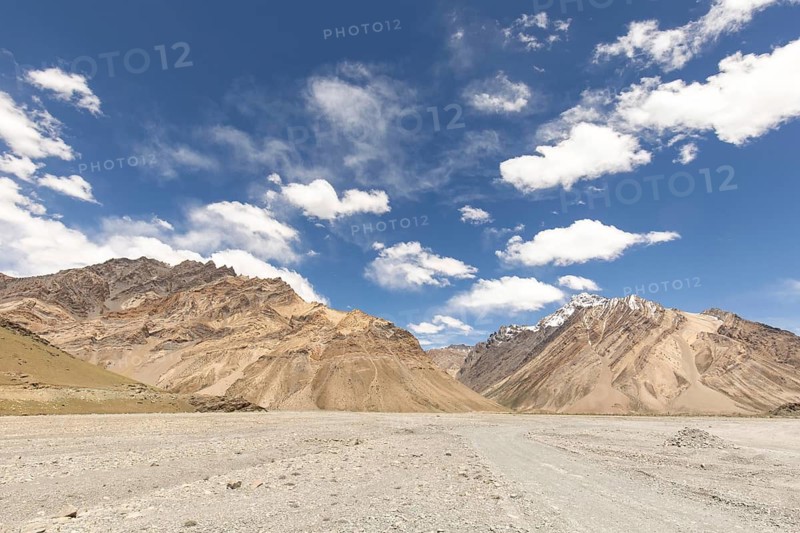 Majestic mountains rise under a clear blue sky, captured in the stunning Zanskar Valley of Ladakh, India. The dry, rocky terrain contrasts beautifully with the cloud dotted sky