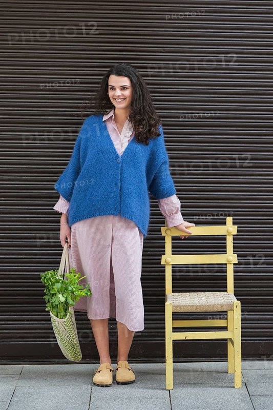 A smiling woman stands next to a yellow chair, wearing a blue cardigan and holding a bag with vegetables. She is outdoors in front of a dark shutter backdrop