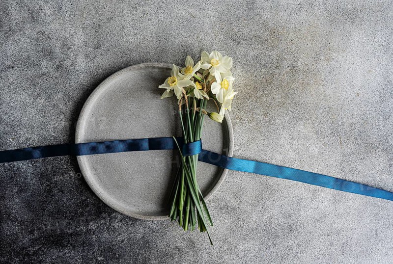 Top view of a bouquet of white daffodils, tied neatly with a dark blue ribbon, resting on a circular gray plate against a textured gray background. The flowers add a touch of elegance and simplicity