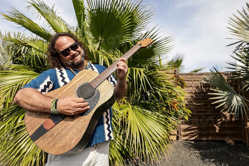 A cheerful man plays a guitar in a sunny, tropical garden Wearing sunglasses and a patterned shirt, he exudes a relaxed, joyful vibe, perfect for a fun, carefree day