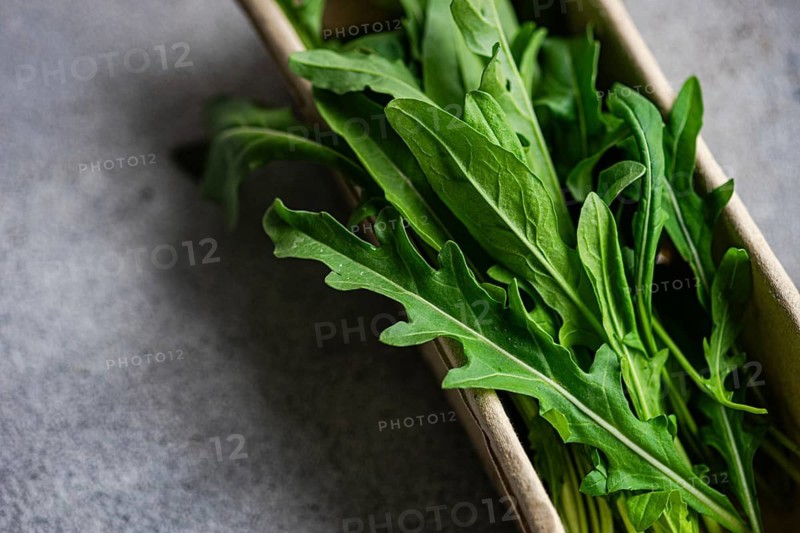 Close up of fresh, vibrant green arugula leaves stacked in a cardboard box. The leaves are crisp and healthy, set against a textured gray background for contrast