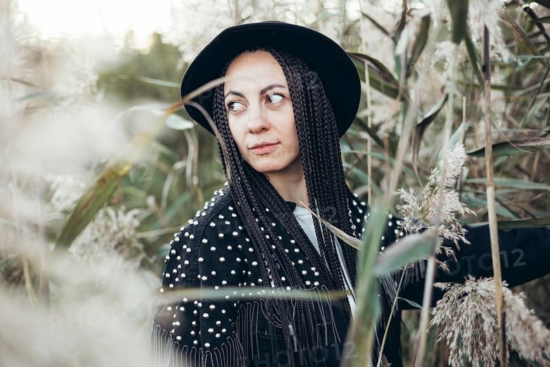A woman stands amidst tall grasses at sunset, her braided hair gently catching the last light of the day. Her black hat adds a touch of elegance to the serene natural setting