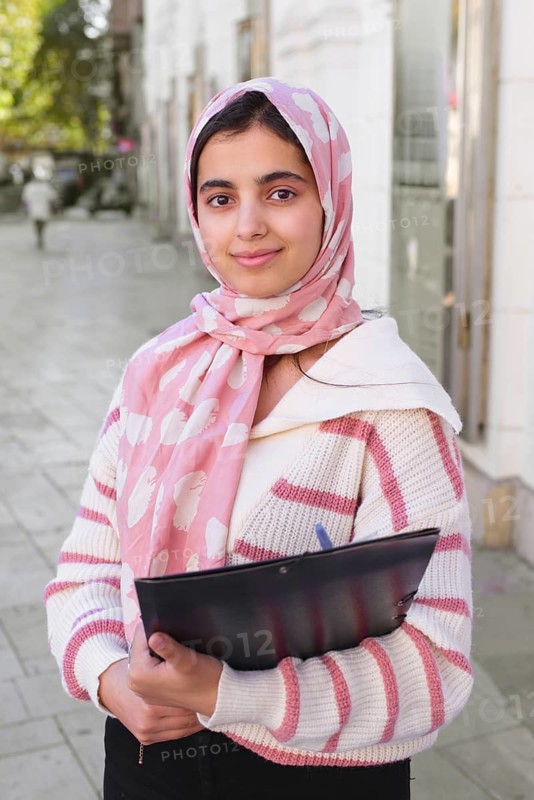 A arab student woman wearing a pink hijab, holding a folder while standing on a city sidewalk. The image captures her friendly expression and professional demeanor