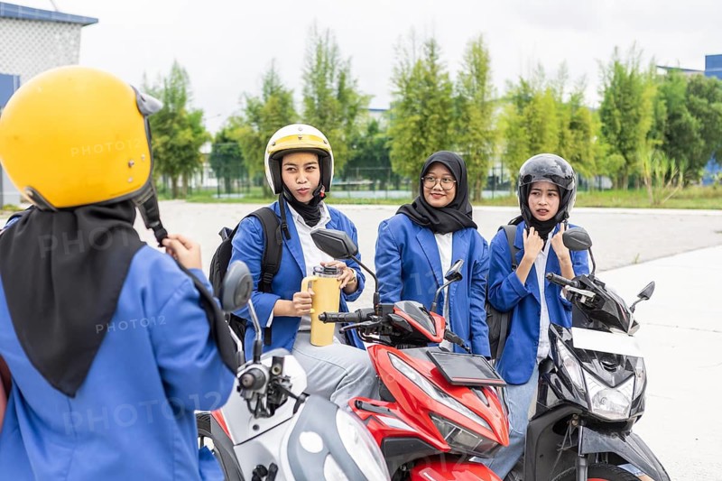 A group of Asian Muslim students wearing helmets and backpacks gather around their motorbikes. They are preparing to ride to school, showcasing a blend of tradition and modernity