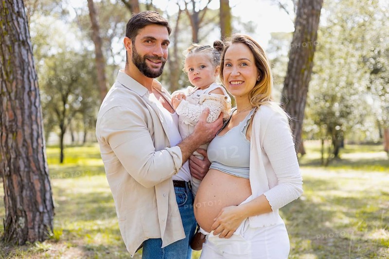A pregnant woman stands with her partner and young child in a sunlit park. They are smiling and enjoying the day together, surrounded by tall trees and greenery