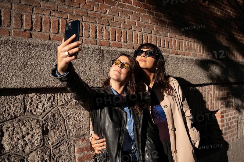 A multiethnic lesbian couple poses for a joyful outdoor selfie, exuding pride and love. The duo stands close, capturing a moment of happiness and unity