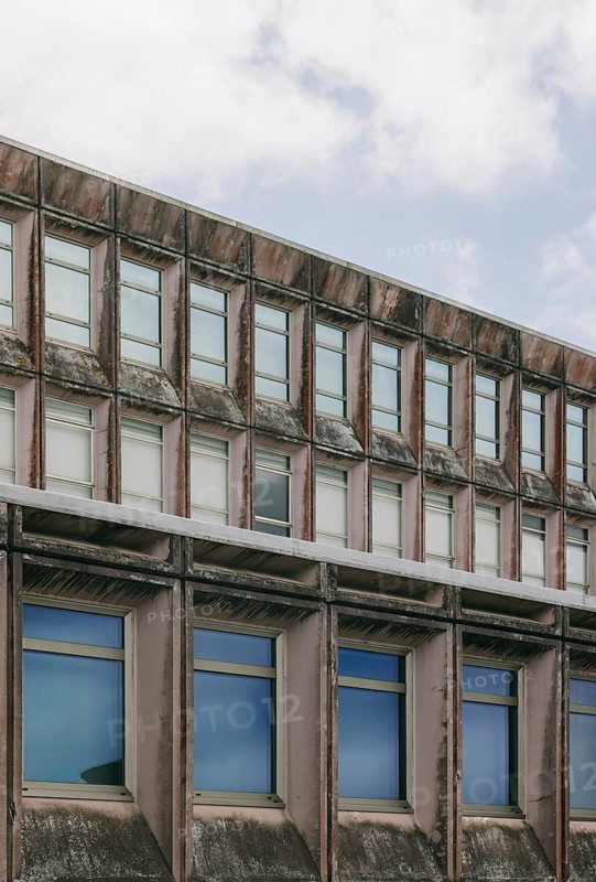 Close-up shot of a weathered brutalist architecture building facade, featuring rows of windows against a cloudy sky. The aged concrete structure displays a raw, rugged appearance