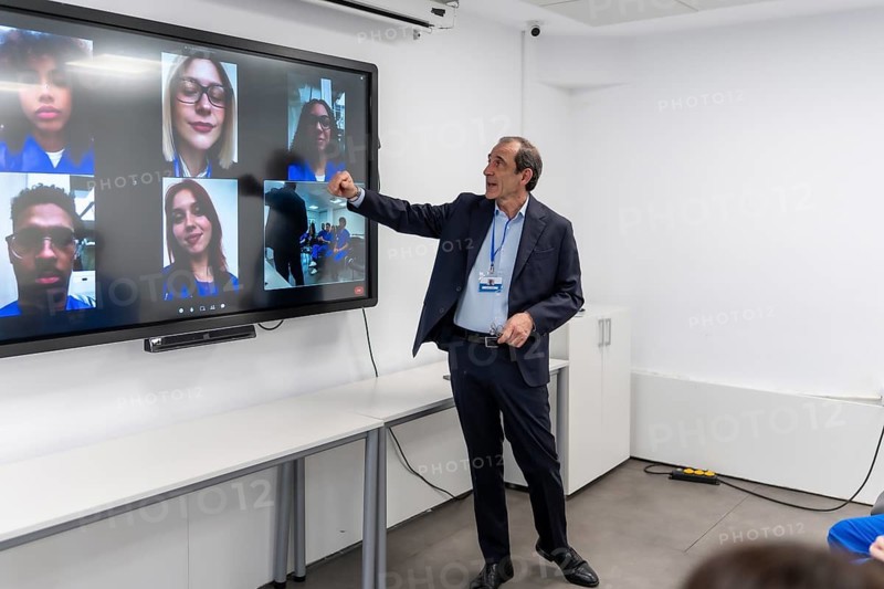 Teacher leads virtual class, pointing to large interactive screen displaying a grid of remote students in a video conference, demonstrating modern digital learning and collaboration
