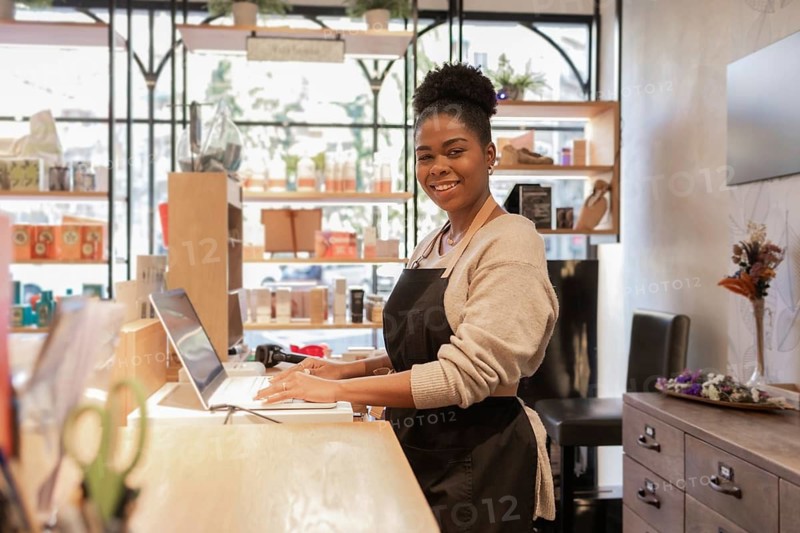 A cheerful store worker wearing a black apron smiles while assisting customers at a cosmetics store, featuring shelves of beauty products and a warm, welcoming atmosphere