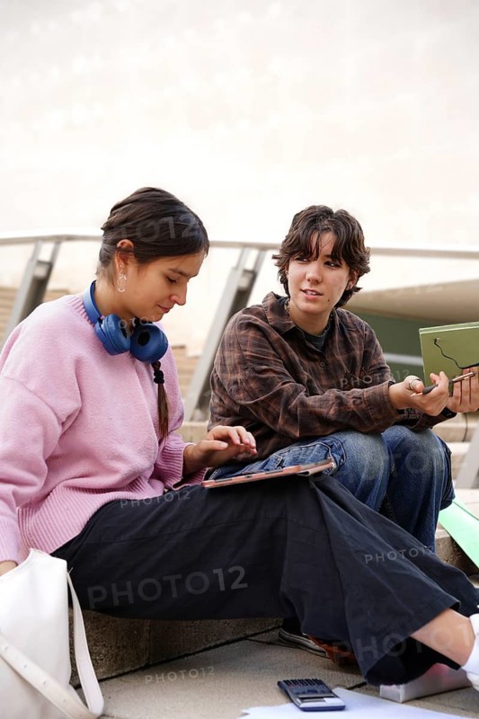 Two teenagers, a non-binary individual and a female, are seen engaging in educational activities outside, with one using a tablet and the other discussing a topic. They sit comfortably on steps, showcasing a relaxed academic environment