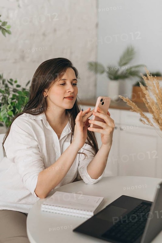 A nutritionist in a modern office setting uses her smartphone for virtual consultations. A laptop and notebook are visible on the table, adding to the professional ambiance
