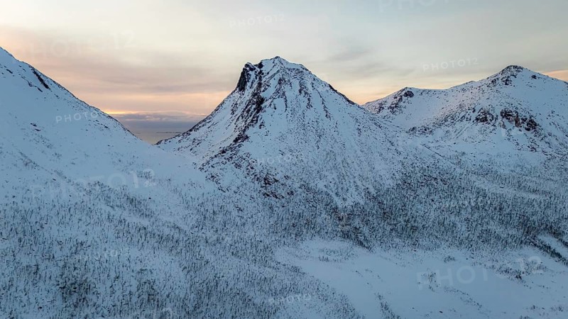 A breathtaking aerial view captures the majestic snow-covered mountains of a Nordic landscape, bathed in soft evening light. The serene vista evokes tranquility and natural beauty, Kvaløya, Troms, Norway