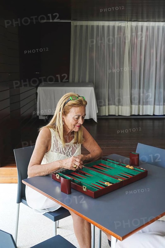 A woman thoughtfully engages in a game of backgammon at a table indoors The board's vibrant colors contrast with the serene setting, conveying focus and strategy in the moment