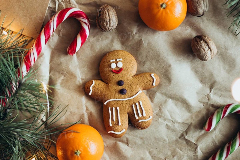 A cheerful gingerbread man cookie decorated with icing, surrounded by candy canes, walnuts, oranges, and pine branches, creating a cozy, festive holiday atmosphere
