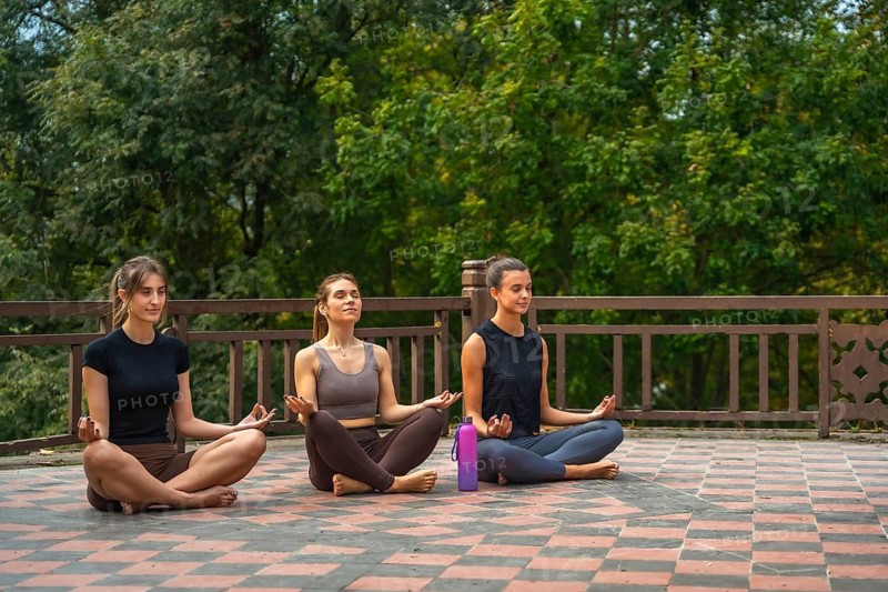 Young women are practicing meditation and yoga together in a serene outdoor setting, sitting cross legged with closed eyes, finding peace and wellness while connecting with nature