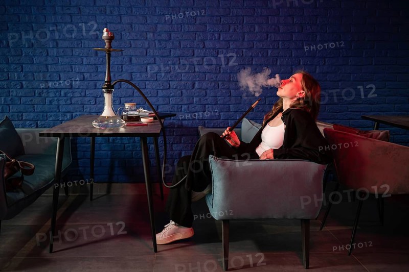 A young woman lounges comfortably in a modern hookah bar, enjoying a session. The space features a stylish setting with a blue brick wall background, creating a relaxed ambiance
