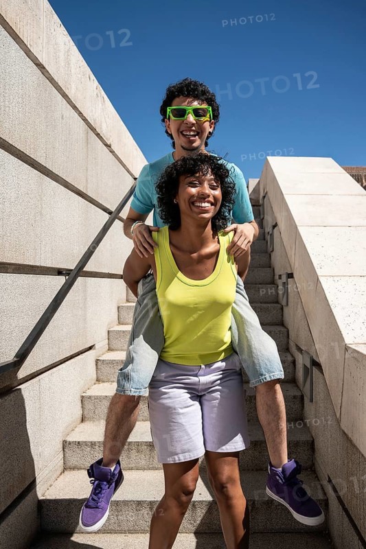 A cheerful multiethnic LGBTQ+ couple has fun outdoors, with one partner playfully giving the other a piggyback ride on sunny steps, showcasing happiness and love