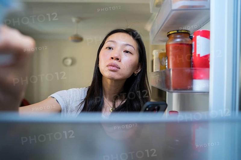 A woman stands in front of an open fridge, using her smartphone to shop for groceries online. The scene highlights technology in daily life and convenient digital shopping