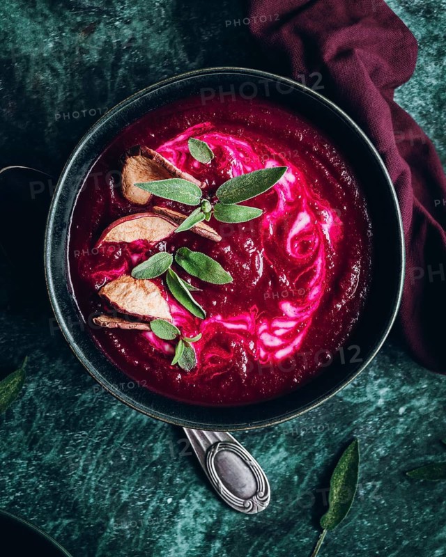 Overhead view of a beetroot soup garnished with slices of apple and fresh herbs. The rich, deep red color contrasts beautifully with the green and wooden tones, creating a visually appealing dish