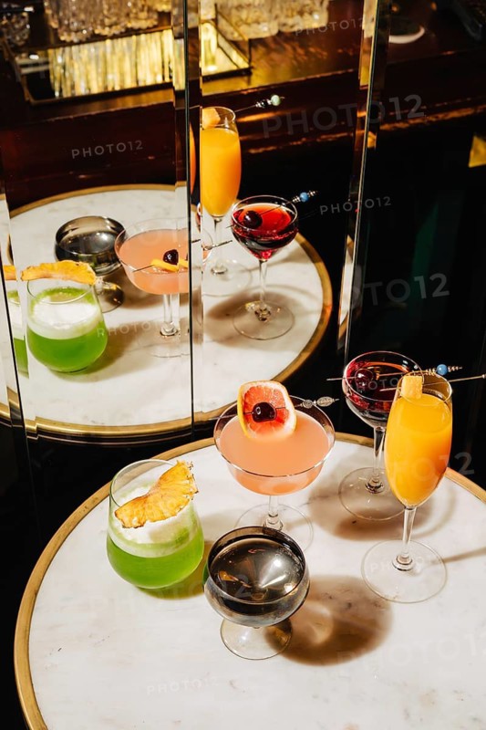 A luxurious selection of mixed cocktails with vibrant colors and unique garnishes, displayed on a marble table. The mirrored reflection enhances the stylish bar ambiance and upscale presentation