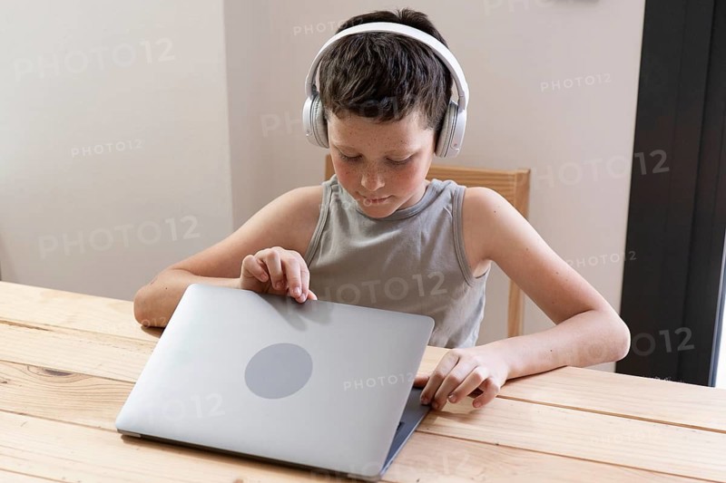 A young child is focused on using a laptop with headphones on at a wooden desk. The setting suggests a home environment, perfect for education or entertainment