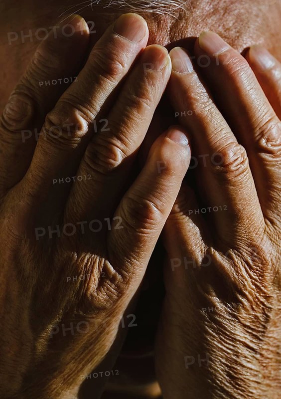 Close-up image of a senior man covering his face with both hands in deep thought The lighting highlights the texture of his skin, conveying emotion and contemplation