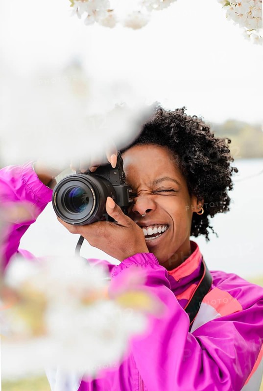 A joyful African American woman photographer, dressed in a colorful jacket, captures the beauty of cherry blossoms with her camera. The petals frame her face, enhancing the scene