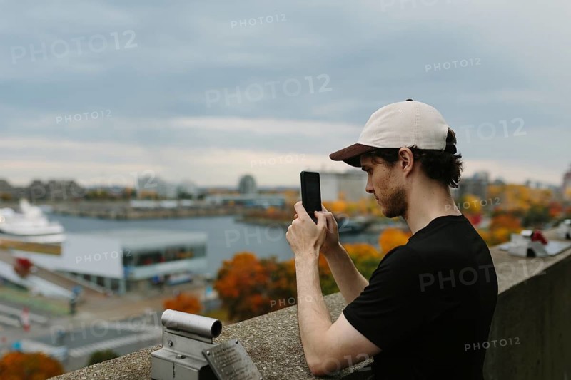 Side view of man in a cap captures a picturesque autumn view of Montreal with his phone, standing by a waterfront overlooking vibrant fall foliage and urban architecture