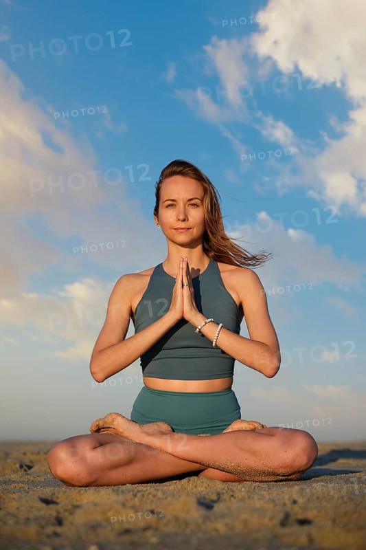 A woman sits in padmasana lotus pose on the beach during sunset, hands in namaste, embodying peace and tranquility amidst nature