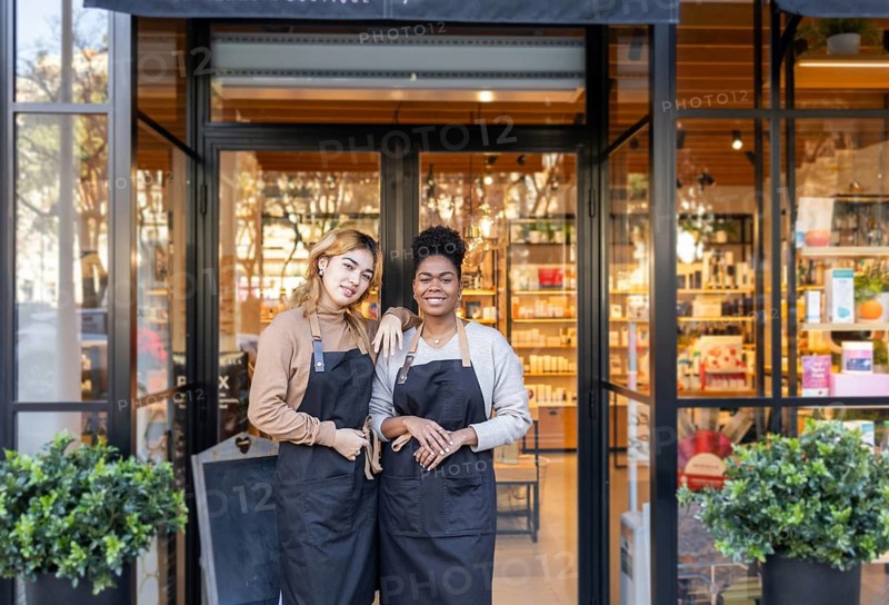 Two smiling employees in aprons stand confidently at the entrance of a cosmetics store. The background showcases shelves with beauty products, creating a welcoming atmosphere