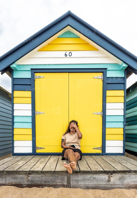 A cheerful woman sits barefoot in front of a colorful Brighton bathing box with yellow doors and striped walls, enjoying a relaxed moment on the beach in Melbourne, Australia