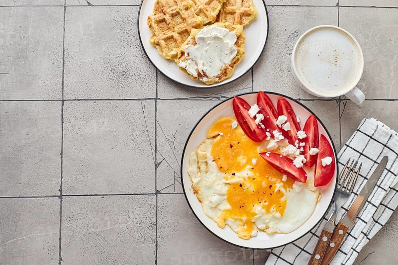 Top view of delectable homemade brunch featuring fried eggs topped with feta cheese and tomatoes, alongside Belgian waffles with cream cheese, accompanied by a fresh cup of coffee