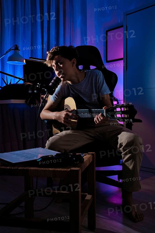 A teenage boy sits on a chair in a dimly lit room, passionately singing and playing an acoustic guitar. The ambient lighting creates a dramatic and intimate atmosphere