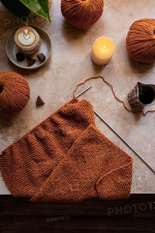 A cozy knitting setup featuring warm brown yarn balls, a knitting project, and a steaming cup of coffee with chocolates, illuminated by a soft candlelight glow