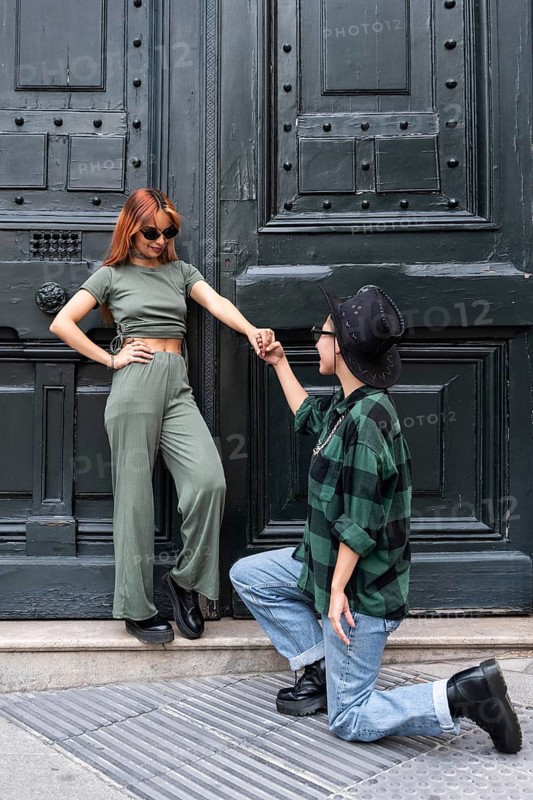 A Colombian lesbian couple shares a romantic moment in front of an elegant dark green door. One partner kneels while holding the other's hand, creating a stylish and affectionate scene