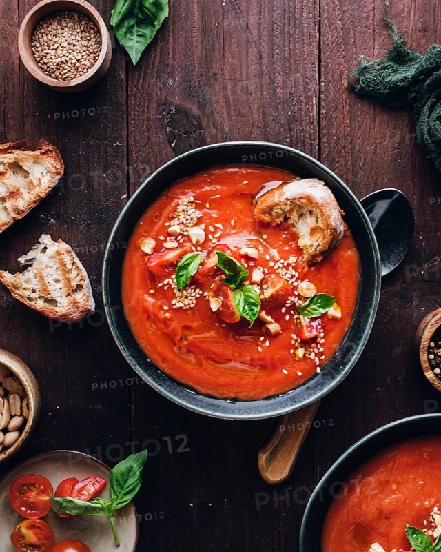 A bowl of vibrant tomato soup garnished with fresh basil leaves and sesame seeds is served on a rustic wooden table, accompanied by crusty bread pieces, creating a cozy dining setup