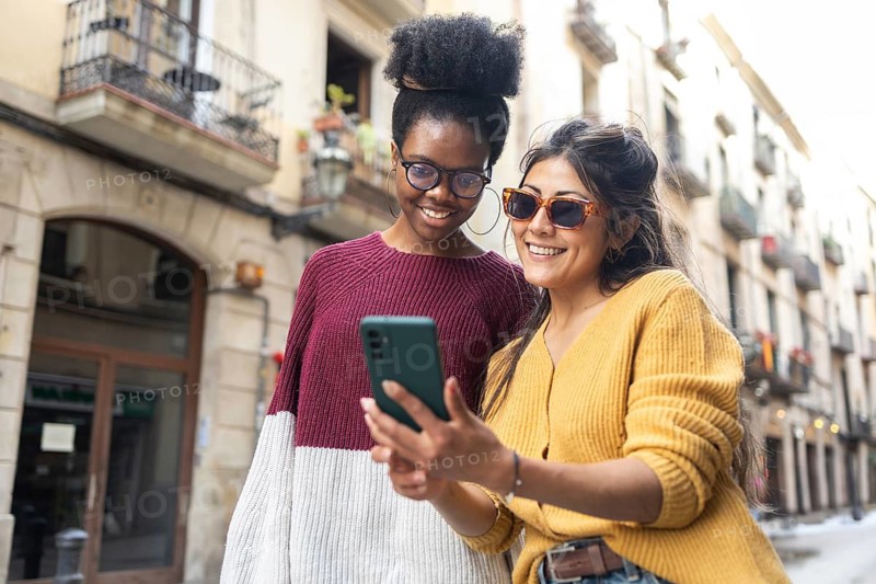Two friends are enjoying a day out in the city. They are smiling and wearing stylish sunglasses while looking at a smartphone. The background features charming architecture