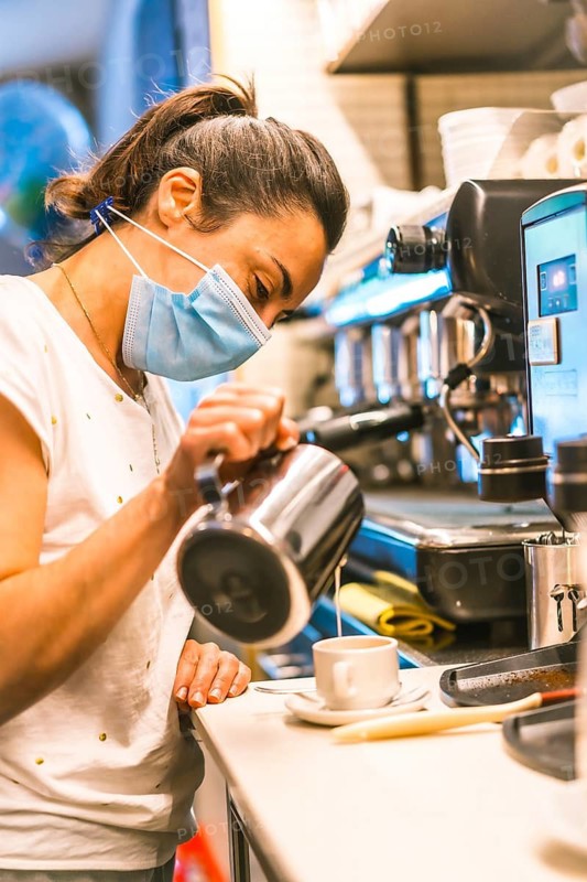 Photo session with a waitress with a face mask in a bar. New normality, security measures after the coronavirus pandemic, social distance, covid-19. Throwing milk into a cup of coffee