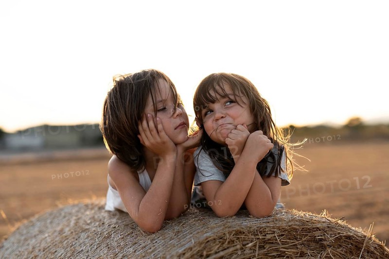 Two children lie on a hay bale in a sunlit field, enjoying each other's company. The warm golden light captures a moment of sibling joy and carefree summer leisure