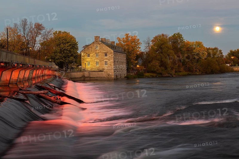 Illuminated Moulin Neuf water flow control dam and walkway over Des Mille-Iles river plus New Mill on Ile des Moulins in autumn at dusk, Old Terrebonne, Quebec, Canada