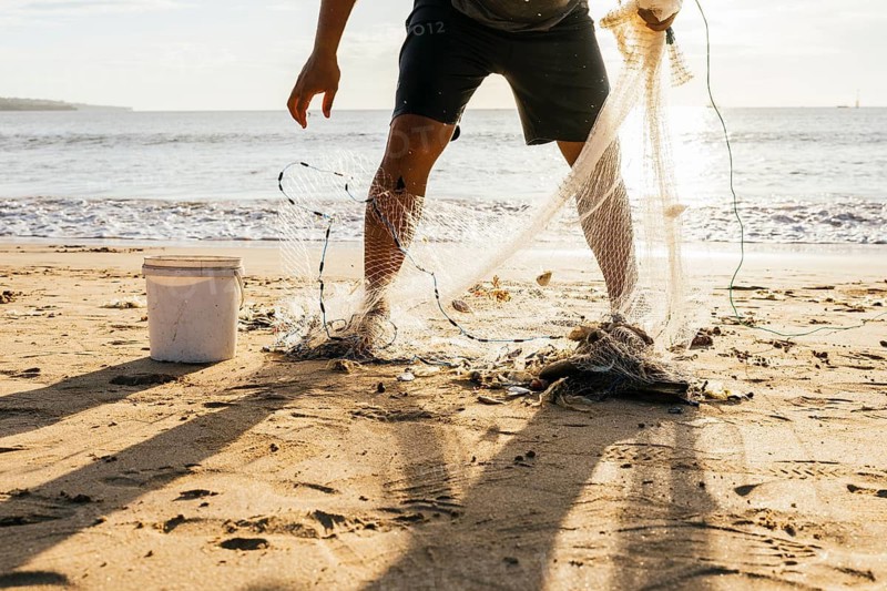 A fisherman stands on a sunlit beach, collecting his catch from a fishing net. Shadows stretch across the sand, and the ocean waves gently lap the shore in the background