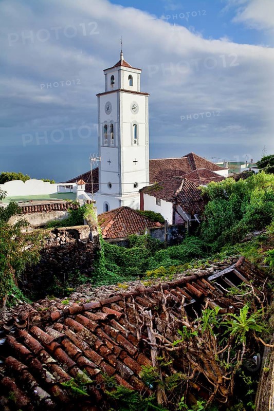 El Tanque showcases its charm with a historic church tower rising above traditional tile roofs and lush greenery, captured under a bright blue sky