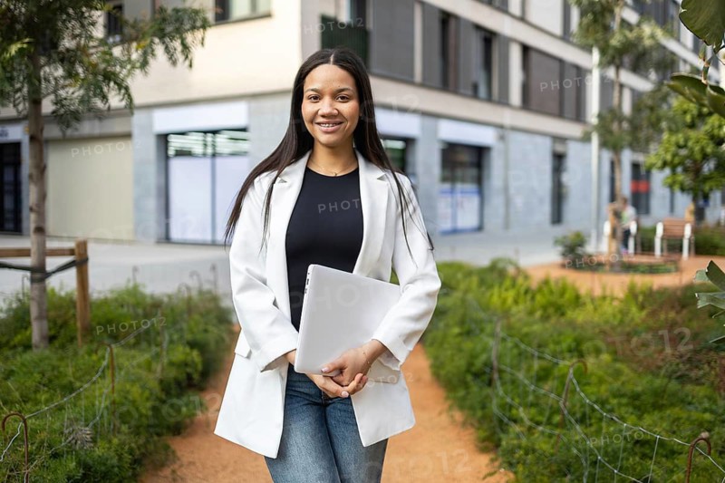 A confident businesswoman in a white blazer carrying a laptop walks through an urban outdoor setting. She exudes professionalism and modern business lifestyle