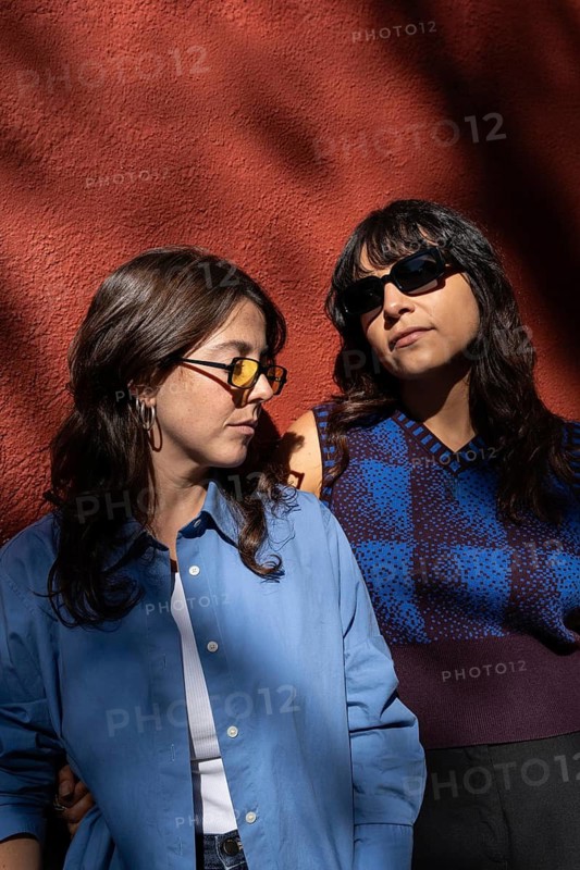 A multiethnic lesbian couple stands together, embodying pride and unity against a vibrant backdrop. Their expressions reflect diversity, strength, and love