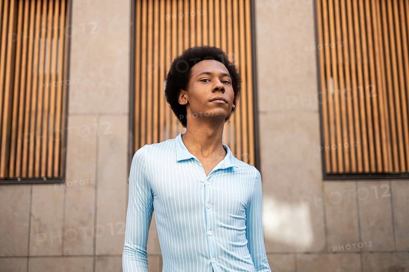 A confident non-binary person stands outdoors, showcasing their identity with pride. The background features a cityscape with modern architecture and wooden panels