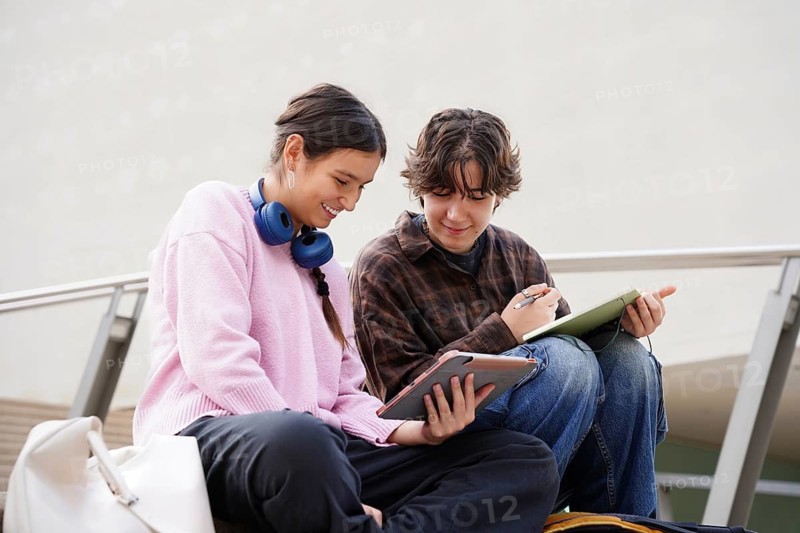 A non-binary teen and a female student sit outdoors, engaging with digital tablets. They share a close bond, reviewing educational content possibly for back-to-school