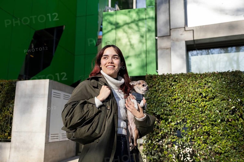 A woman in a green jacket holds a small chihuahua while enjoying a sunny day outside a modern building with green panels and lush greenery in the background