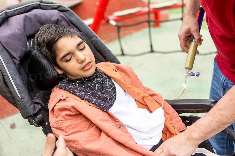 A child with cerebral palsy in a wheelchair receives attentive care in a playground setting. The scene reflects support, and care with emphasis on health and personal well being
