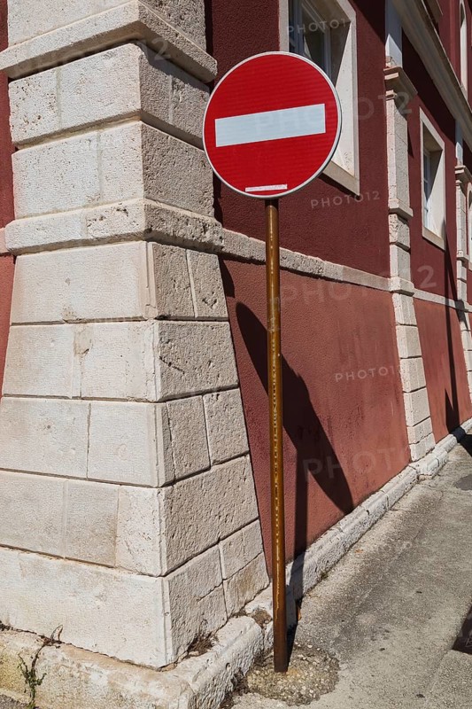 Red and white circular Do Not Enter pictogram notice sign for motor vehicle circulation attached to an old rusted steel post on sidewalk next to an unusual architectural style building at a street corner, Zadar, Croatia