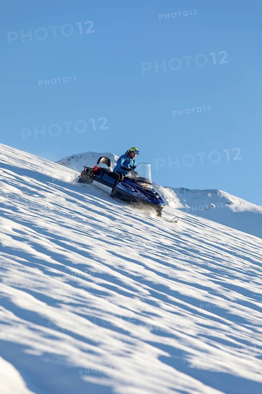 A person on a snowmobile speeds across a pristine, snowy slope with clear blue skies. Surrounded by winter mountains, the scene captures the thrill of outdoor adventure